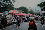 A busy street market with numerous vendors under colorful tents and umbrellas. People are gathered around stalls, some wearing masks. Motorbikes are parked nearby, and trees and signboards are visible in the background.