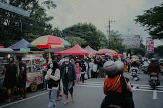 A busy street market with numerous vendors under colorful tents and umbrellas. People are gathered around stalls, some wearing masks. Motorbikes are parked nearby, and trees and signboards are visible in the background.