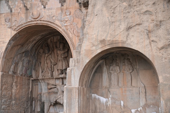 Stone carvings are set within arched recesses of an ancient structure. Intricately detailed figures, including human-like forms and angels, are etched into the stone, possibly depicting historical or mythological scenes. The surface features a weathered texture with natural rock hues.