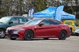 A red sports sedan is parked in an outdoor setting, with promotional tents and banners in the background. The vehicle's sleek design and black wheels are prominent, and it is parked alongside other cars of different colors.