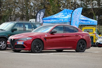 A red sports sedan is parked in an outdoor setting, with promotional tents and banners in the background. The vehicle's sleek design and black wheels are prominent, and it is parked alongside other cars of different colors.