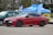 A red sports sedan is parked in an outdoor setting, with promotional tents and banners in the background. The vehicle's sleek design and black wheels are prominent, and it is parked alongside other cars of different colors.