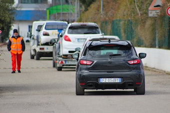 Several cars are lined up on a road, with a person wearing a bright orange safety vest and red pants standing to the side. The rear of a black car with a visible license plate is in the foreground, followed by another car on a trailer and additional vehicles in a row. The setting appears to be a road or checkpoint area, with slight greenery visible on the side.