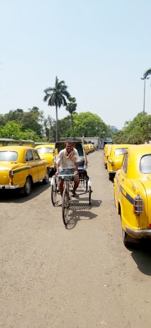 A street scene with yellow taxis parked on either side. A man is riding a bicycle rickshaw down the middle of the road. The surroundings include palm trees and greenery under a clear blue sky.