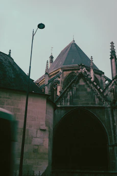 A gothic-style church with intricate architectural details, including pointed arches and a steep roof. A streetlamp stands nearby, silhouetted against the sky, adding a touch of modernity to the historical structure. The image has a muted, moody color palette.