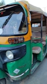 An auto rickshaw with a yellow and green color scheme, featuring a partially open side, is parked on the road. The vehicle appears slightly worn with visible signs of use, and it has a CNG sticker on the front.