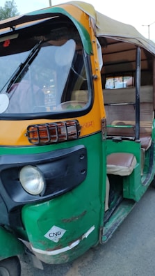 An auto rickshaw with a yellow and green color scheme, featuring a partially open side, is parked on the road. The vehicle appears slightly worn with visible signs of use, and it has a CNG sticker on the front.
