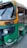 An auto rickshaw with a yellow and green color scheme, featuring a partially open side, is parked on the road. The vehicle appears slightly worn with visible signs of use, and it has a CNG sticker on the front.