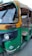 An auto rickshaw with a yellow and green color scheme, featuring a partially open side, is parked on the road. The vehicle appears slightly worn with visible signs of use, and it has a CNG sticker on the front.
