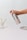 A hand is holding a metal spray bottle labeled 'KINN' against a minimalist background with small dark specks. On the table, there is a glass cup containing two toothbrushes with wooden handles and a white ceramic soap dish.