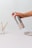 A hand is holding a metal spray bottle labeled 'KINN' against a minimalist background with small dark specks. On the table, there is a glass cup containing two toothbrushes with wooden handles and a white ceramic soap dish.
