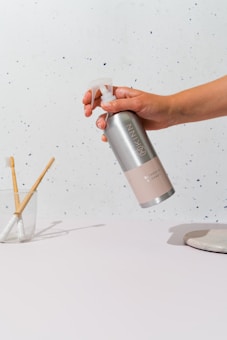 A hand is holding a metal spray bottle labeled 'KINN' against a minimalist background with small dark specks. On the table, there is a glass cup containing two toothbrushes with wooden handles and a white ceramic soap dish.