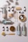 A collection of eco-friendly cleaning products and personal care items laid out on a flat surface. The composition includes bottles of cleaning sprays with silver metallic finishes, glass bottles of essential oils, wooden toothbrushes, a rectangular bar of natural soap, and various cleaning brushes with wooden handles. The arrangement is visually appealing, with shadows adding depth and texture.