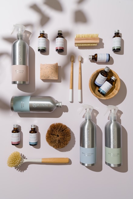 A collection of eco-friendly cleaning products and personal care items laid out on a flat surface. The composition includes bottles of cleaning sprays with silver metallic finishes, glass bottles of essential oils, wooden toothbrushes, a rectangular bar of natural soap, and various cleaning brushes with wooden handles. The arrangement is visually appealing, with shadows adding depth and texture.