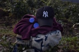 A dark-colored baseball cap with a white embroidered logo is placed on top of a folded maroon hoodie. Next to the hoodie is a pair of round, blue-tinted sunglasses. These items are resting on a blue and white tie-dye patterned bag with zippers. The background consists of green foliage and plants, suggesting an outdoor setting.