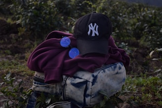 A dark-colored baseball cap with a white embroidered logo is placed on top of a folded maroon hoodie. Next to the hoodie is a pair of round, blue-tinted sunglasses. These items are resting on a blue and white tie-dye patterned bag with zippers. The background consists of green foliage and plants, suggesting an outdoor setting.