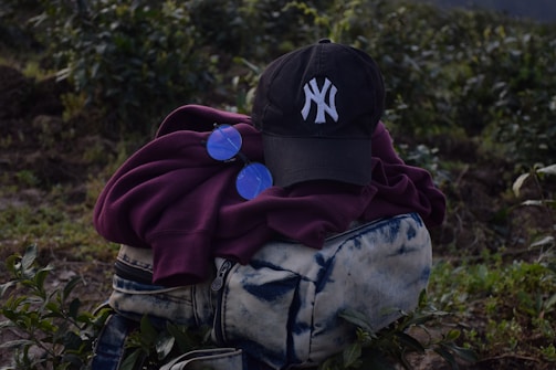 A dark-colored baseball cap with a white embroidered logo is placed on top of a folded maroon hoodie. Next to the hoodie is a pair of round, blue-tinted sunglasses. These items are resting on a blue and white tie-dye patterned bag with zippers. The background consists of green foliage and plants, suggesting an outdoor setting.
