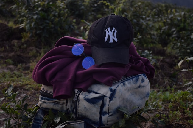 A dark-colored baseball cap with a white embroidered logo is placed on top of a folded maroon hoodie. Next to the hoodie is a pair of round, blue-tinted sunglasses. These items are resting on a blue and white tie-dye patterned bag with zippers. The background consists of green foliage and plants, suggesting an outdoor setting.