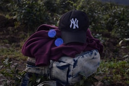 A dark-colored baseball cap with a white embroidered logo is placed on top of a folded maroon hoodie. Next to the hoodie is a pair of round, blue-tinted sunglasses. These items are resting on a blue and white tie-dye patterned bag with zippers. The background consists of green foliage and plants, suggesting an outdoor setting.