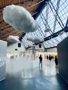Inside a large indoor space with a high, glass-paneled ceiling, several fluffy artificial clouds are suspended. A ladder leads up to one of the clouds, creating a surreal atmosphere. There are people walking and observing various artworks displayed on the walls of what appears to be an art gallery or exhibition.