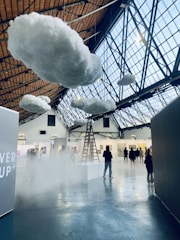 Inside a large indoor space with a high, glass-paneled ceiling, several fluffy artificial clouds are suspended. A ladder leads up to one of the clouds, creating a surreal atmosphere. There are people walking and observing various artworks displayed on the walls of what appears to be an art gallery or exhibition.