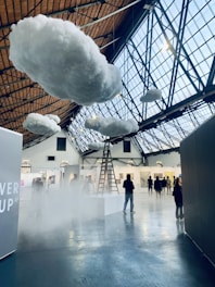 Inside a large indoor space with a high, glass-paneled ceiling, several fluffy artificial clouds are suspended. A ladder leads up to one of the clouds, creating a surreal atmosphere. There are people walking and observing various artworks displayed on the walls of what appears to be an art gallery or exhibition.