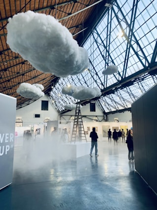 Inside a large indoor space with a high, glass-paneled ceiling, several fluffy artificial clouds are suspended. A ladder leads up to one of the clouds, creating a surreal atmosphere. There are people walking and observing various artworks displayed on the walls of what appears to be an art gallery or exhibition.