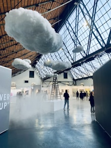 Inside a large indoor space with a high, glass-paneled ceiling, several fluffy artificial clouds are suspended. A ladder leads up to one of the clouds, creating a surreal atmosphere. There are people walking and observing various artworks displayed on the walls of what appears to be an art gallery or exhibition.