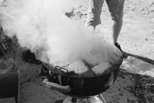A barbecue grill is in use, producing a significant amount of smoke. Several pieces of food, possibly meat, are visible on the grill. The background suggests an outdoor setting with someone standing nearby wearing shorts and sneakers.
