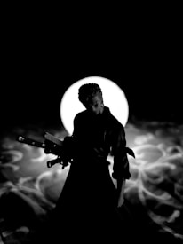 A silhouetted figure stands prominently against a backdrop of a bright, circular light. The character holds two swords and is likely a warrior or samurai, depicted in an action-ready pose. The background features swirling patterns reminiscent of smoke or mist.