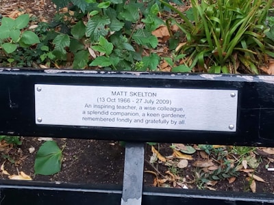 A memorial bench plaque dedicated to Matt Skelton, set against a background of green foliage. The plaque mentions his dates of birth and death, and honors him as an inspiring teacher, wise colleague, splendid companion, and keen gardener.