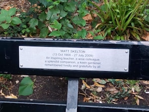A memorial bench plaque dedicated to Matt Skelton, set against a background of green foliage. The plaque mentions his dates of birth and death, and honors him as an inspiring teacher, wise colleague, splendid companion, and keen gardener.