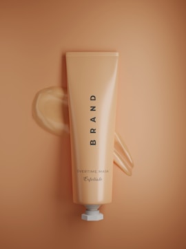 A neatly designed tube with the word 'BRAND' vertically aligned at the center. Below it, the text 'OVERTIME MASK' with 'Exfoliate' written underneath in a cursive font. The tube appears to be a product packaging against a matching peach-colored background with a subtle texture. A small amount of product is visible, squeezed out near the top of the tube.