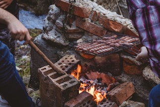 Two people are tending to a barbecue grill over a fire in an outdoor setting. The fire is set within a makeshift pit made of bricks, and sausages are being cooked on a metal grill rack. The area appears natural, possibly near a stream, as water is visible in the background.