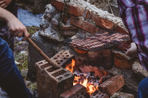 Two people are tending to a barbecue grill over a fire in an outdoor setting. The fire is set within a makeshift pit made of bricks, and sausages are being cooked on a metal grill rack. The area appears natural, possibly near a stream, as water is visible in the background.