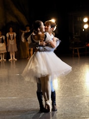 A couple dressed in ballet costumes is performing on stage, with the male dancer lifting the female dancer, who wears a white tutu. They are surrounded by soft lighting, with other dancers in the background wearing period costumes.