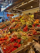 A vibrant market stall overflowing with a variety of fresh fruits and vegetables, including tomatoes, pineapples, peppers, beans, and pumpkins. The produce is artfully arranged in an inviting, colorful display under bright lighting. Shoppers are visible in the background, adding a lively atmosphere to the scene.