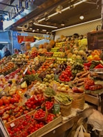 A vibrant market stall overflowing with a variety of fresh fruits and vegetables, including tomatoes, pineapples, peppers, beans, and pumpkins. The produce is artfully arranged in an inviting, colorful display under bright lighting. Shoppers are visible in the background, adding a lively atmosphere to the scene.