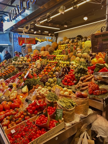 A vibrant market stall overflowing with a variety of fresh fruits and vegetables, including tomatoes, pineapples, peppers, beans, and pumpkins. The produce is artfully arranged in an inviting, colorful display under bright lighting. Shoppers are visible in the background, adding a lively atmosphere to the scene.