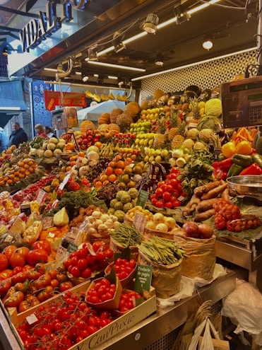 A vibrant market stall overflowing with a variety of fresh fruits and vegetables, including tomatoes, pineapples, peppers, beans, and pumpkins. The produce is artfully arranged in an inviting, colorful display under bright lighting. Shoppers are visible in the background, adding a lively atmosphere to the scene.