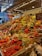 A vibrant market stall overflowing with a variety of fresh fruits and vegetables, including tomatoes, pineapples, peppers, beans, and pumpkins. The produce is artfully arranged in an inviting, colorful display under bright lighting. Shoppers are visible in the background, adding a lively atmosphere to the scene.