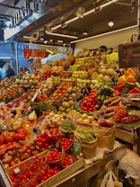 A vibrant market stall overflowing with a variety of fresh fruits and vegetables, including tomatoes, pineapples, peppers, beans, and pumpkins. The produce is artfully arranged in an inviting, colorful display under bright lighting. Shoppers are visible in the background, adding a lively atmosphere to the scene.