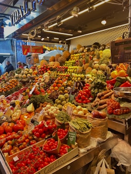 A vibrant market stall overflowing with a variety of fresh fruits and vegetables, including tomatoes, pineapples, peppers, beans, and pumpkins. The produce is artfully arranged in an inviting, colorful display under bright lighting. Shoppers are visible in the background, adding a lively atmosphere to the scene.