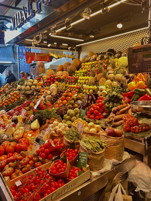 A vibrant market stall overflowing with a variety of fresh fruits and vegetables, including tomatoes, pineapples, peppers, beans, and pumpkins. The produce is artfully arranged in an inviting, colorful display under bright lighting. Shoppers are visible in the background, adding a lively atmosphere to the scene.
