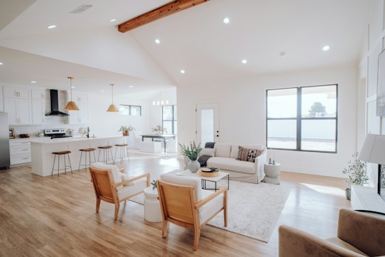 A spacious and bright living area with wooden flooring features a modern open-plan layout. There's a kitchen with white cabinetry and a black range hood in the background. Two pendant lights with wooden accents hang over a long counter with four high stools. The living area includes a white sofa adorned with patterned cushions, a light-colored rug, a coffee table with decorative items, and indoor plants. Natural light floods the space from large windows, enhancing the airy and cozy ambiance.