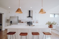A modern kitchen featuring a large white island with brown leather stools. Above the island hang two bronze pendant lights. The cabinetry is white with gold handles, and a stainless steel fridge is visible. A black range hood is centered above a stove, while various kitchen items decorate the countertops. A potted plant decorates the island, and natural light enters through a window.