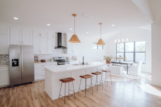 A modern kitchen and dining area featuring white cabinetry and a stainless steel refrigerator. The center island has a white countertop with four brown bar stools. Above the island, two gold pendant lights hang. The dining area has a long table with chairs and is adorned with a plant. The space has large windows providing ample natural light, and the flooring is wooden.