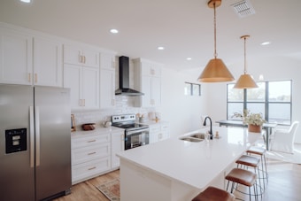 A modern kitchen with white cabinetry and a large island with barstools. The room features stainless steel appliances including a refrigerator and oven. Copper pendant lights hang over the island, which has a sink and a vase of flowers. The space has wooden flooring and large windows allowing natural light to flood the area, creating a bright and airy atmosphere.