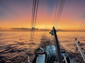 A serene ocean scene at sunset featuring the deck of a sailboat. The sky is filled with warm orange and pink hues, reflecting on the calm waters. The boat's rigging is visible, capturing a peaceful moment on the sea.