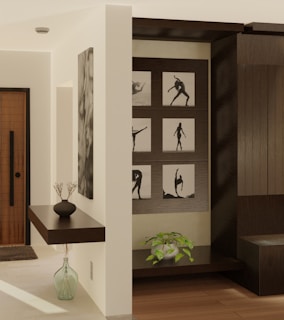 A modern interior with a light wooden floor and cream-colored walls. On the left, a floating dark wooden shelf holds a small black vase with dry twigs. Below the shelf, a larger transparent vase is placed on the floor. The entrance door has a dark wood finish with a vertical handle. Artwork featuring silhouettes of dancers is displayed on a dark wood panel. A small green plant in a white pot adds a touch of nature to the space.